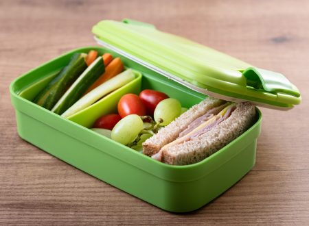 Close-up of a green lunchbox on a wooden table, containing a ham and cheese sandwich, grapes, cherry tomatoes, and vegetable sticks