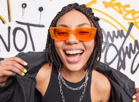 Swag. Fashionable cheerful hipster girl with braids smiles broadly shows white teeth wears trendy orange sunglasses black jacket expresses positive emotions poses against graffiti wall in urban place